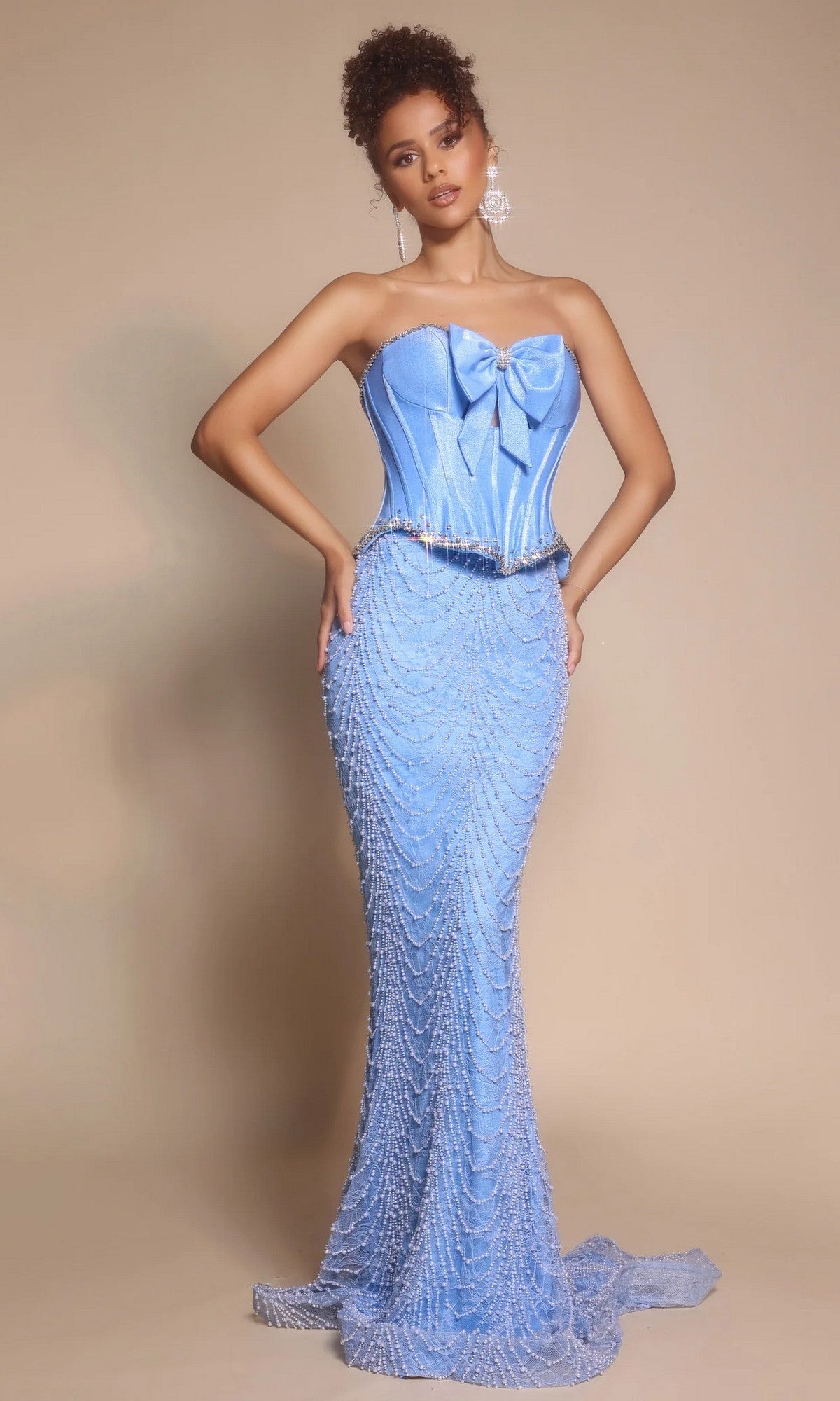 A woman poses against a neutral background in a blue Portia and Scarlett PS26054 long formal dress featuring a fitted, strapless corset bodice, large bow detail, and beaded draping accents.